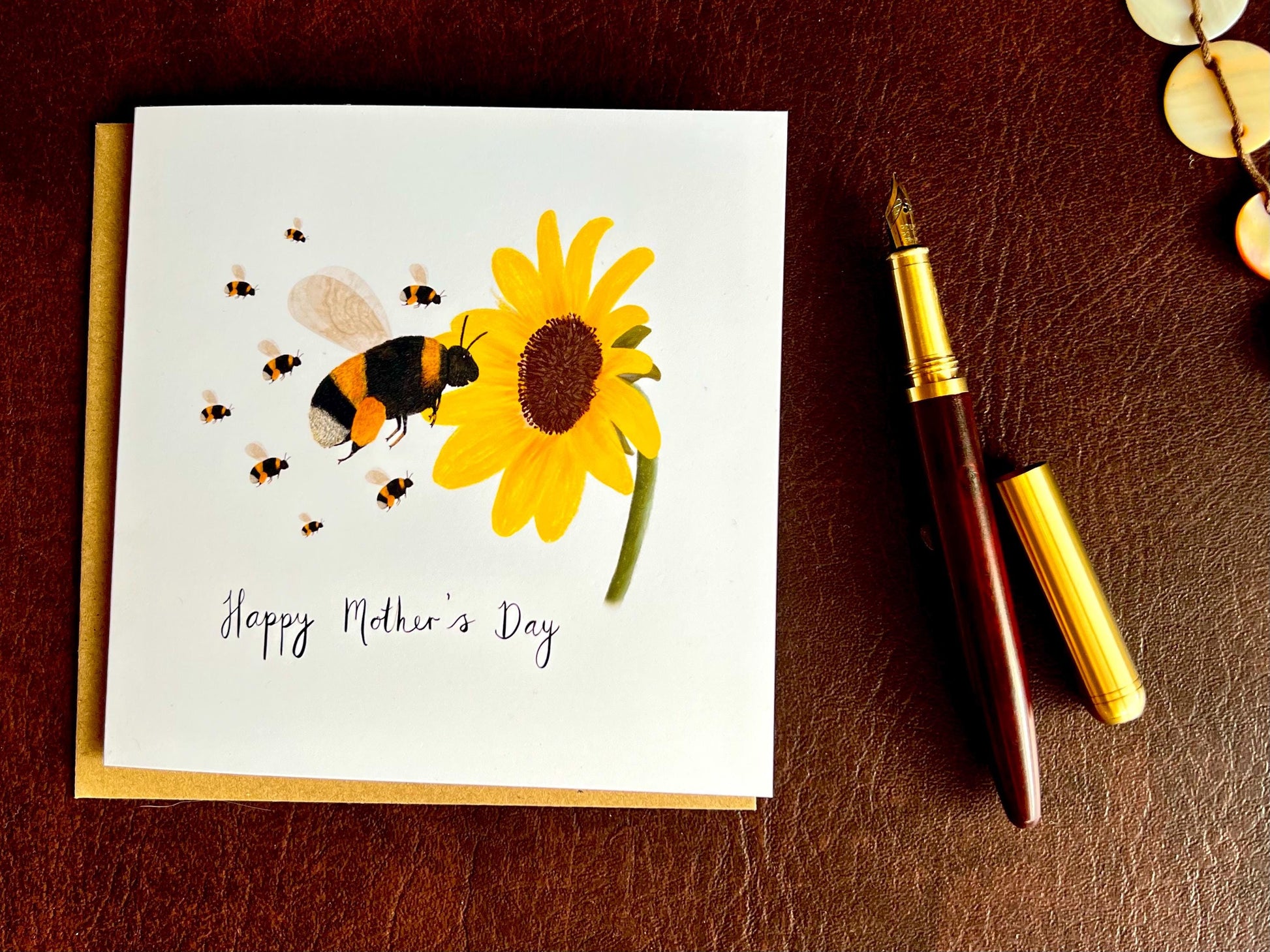 Flat lay image of a handmade Mother’s Day card featuring a bumblebee flying towards a sunflower with a baby bee, shown laid flat on top of a kraft envelope on a brown leatherette tabletop beside a fountain pen.