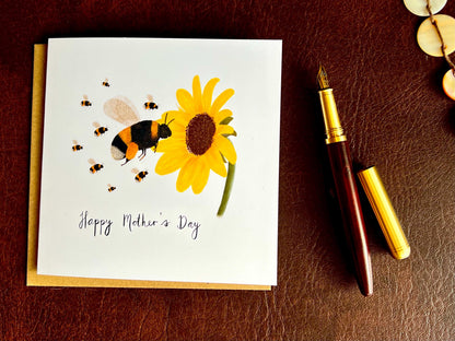 Flat lay image of a handmade Mother’s Day card featuring a bumblebee flying towards a sunflower with a baby bee, shown laid flat on top of a kraft envelope on a brown leatherette tabletop beside a fountain pen.