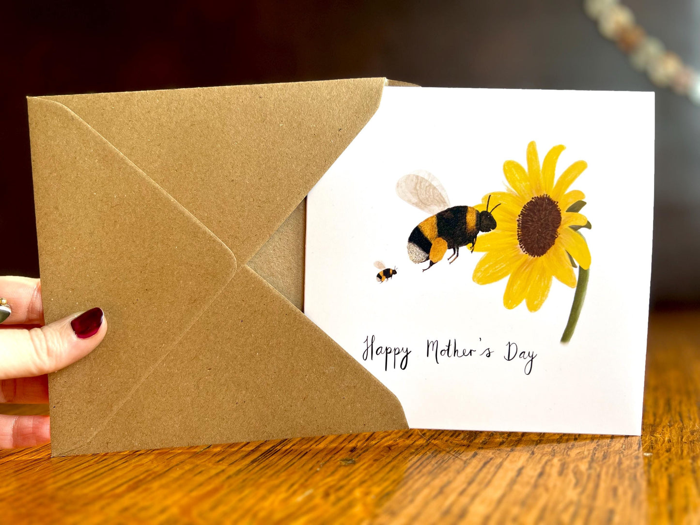 Hand holding a handmade Mother’s Day card being removed from a kraft envelope and held upright towards the camera, featuring an illustrated bumblebee flying towards a sunflower with one baby bee shown as an example of customisation options.