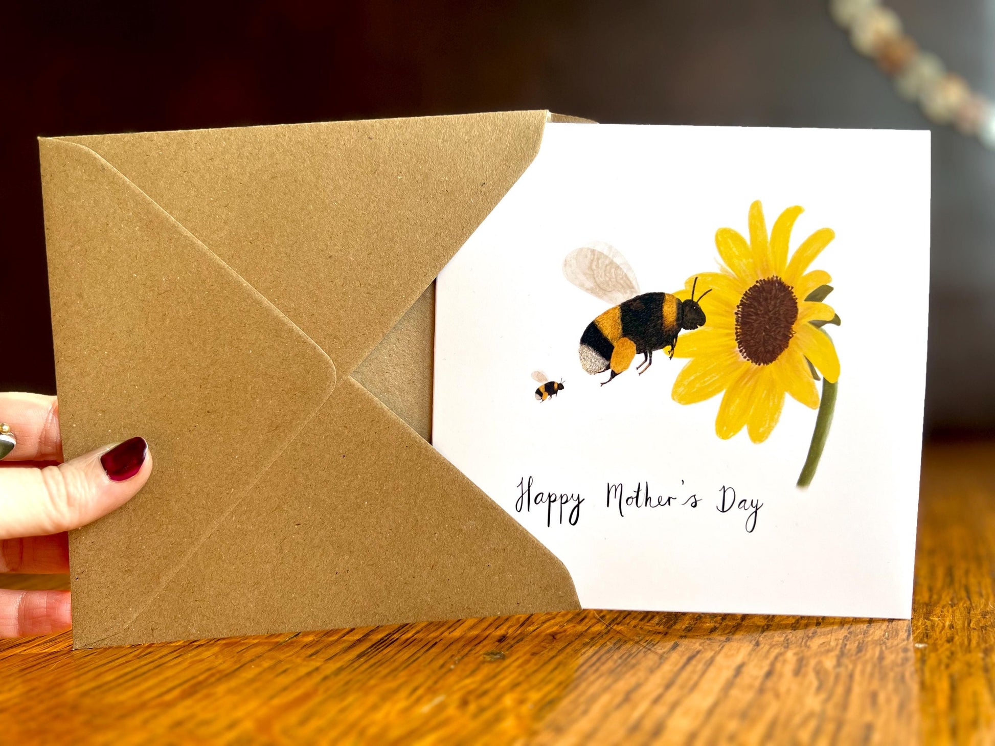 Hand holding a handmade Mother’s Day card being removed from a kraft envelope and held upright towards the camera, featuring an illustrated bumblebee flying towards a sunflower with one baby bee shown as an example of customisation options.