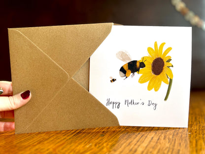 Hand holding a handmade Mother’s Day card being removed from a kraft envelope and held upright towards the camera, featuring an illustrated bumblebee flying towards a sunflower with one baby bee shown as an example of customisation options.