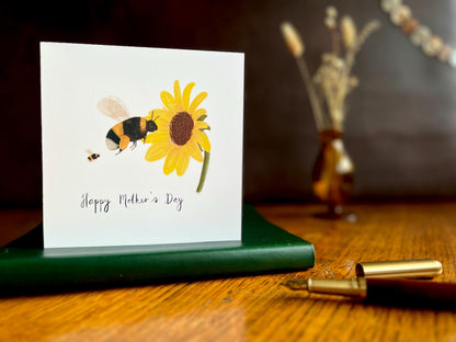Lifestyle image of a handmade Mother’s Day card featuring a bumblebee flying towards a sunflower with a baby bee, displayed standing on a green leatherette book on a wooden tabletop. The scene includes a fountain pen in the foreground, a dark leatherette backdrop, and a blurred vase of dried flowers in the background.