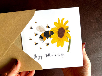 Hands holding a handmade Mother’s Day card being removed from a kraft envelope, featuring an illustrated bumblebee flying towards a yellow sunflower with additional baby bees following behind. The card reads 'Happy Mother’s Day' and can be customised with the number of baby bees to represent children or grandchildren.