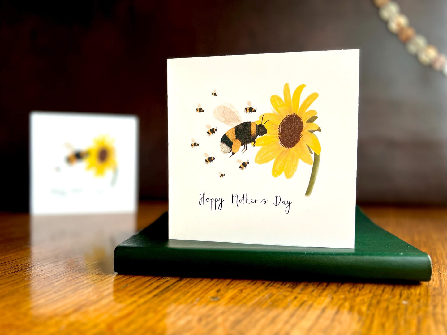 Lifestyle image of a special Mother’s Day card featuring a bumblebee flying towards a sunflower with eight baby bees, displayed standing on a green leatherette book on a tabletop. In the background, a blurred version of the same card shows a single baby bee to demonstrate customisation options.