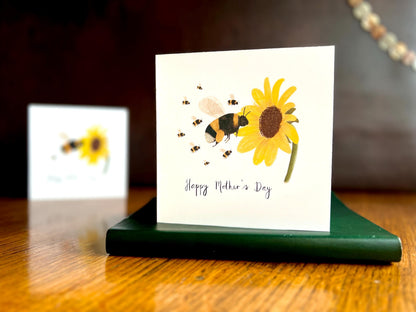 Lifestyle image of a special Mother’s Day card featuring a bumblebee flying towards a sunflower with eight baby bees, displayed standing on a green leatherette book on a tabletop. In the background, a blurred version of the same card shows a single baby bee to demonstrate customisation options.