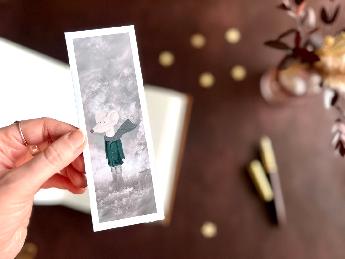 Hand holding a mouse bookmark with white mouse illustration, styled with a fountain pen and open sketchbook in a cosy dark academia inspired desk scene.