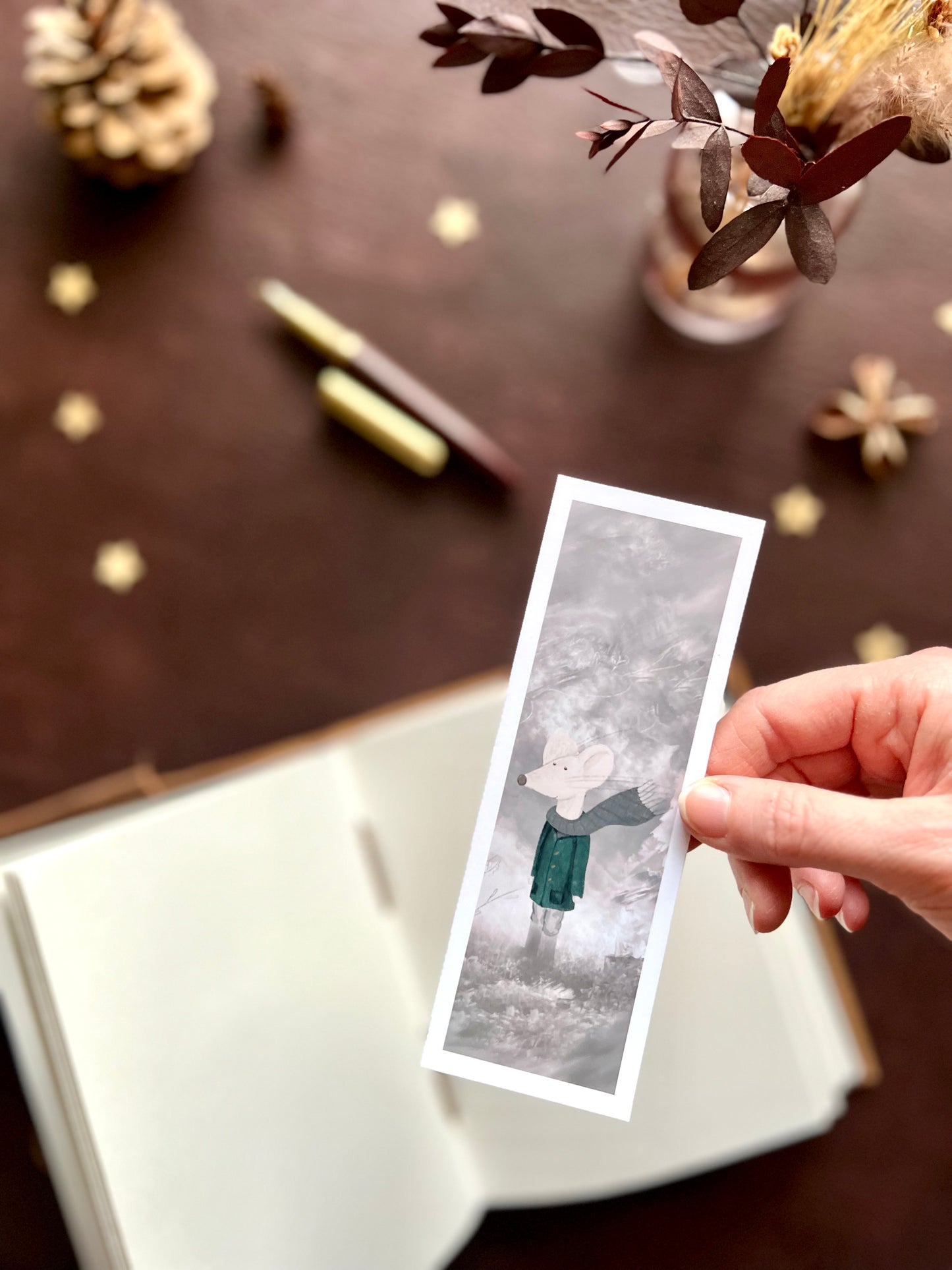 Hand holding a mouse bookmark featuring a white mouse in coat and scarf, with a soft blurred background of pine cones and flowers in a cosy nature-inspired setting.