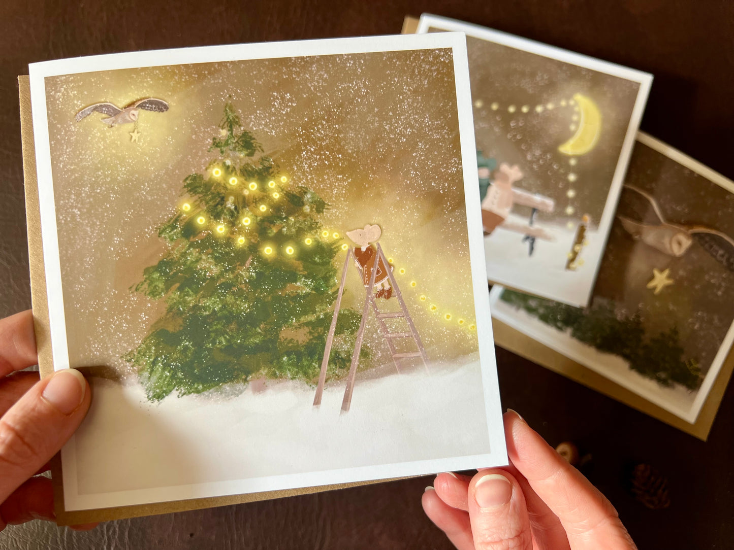 Premium Christmas card from a set of three woodland designs, featuring a storybook-style illustration of a mouse decorating a Christmas tree with fairy lights in a snowy winter scene. The card is shown in close-up being held in two hands, with the other two designs softly out of focus in the background. Ideal for festive gifting, animal lovers and rustic Christmas card sets. Printed on thick 312gsm fine art card by a UK independent artist.
