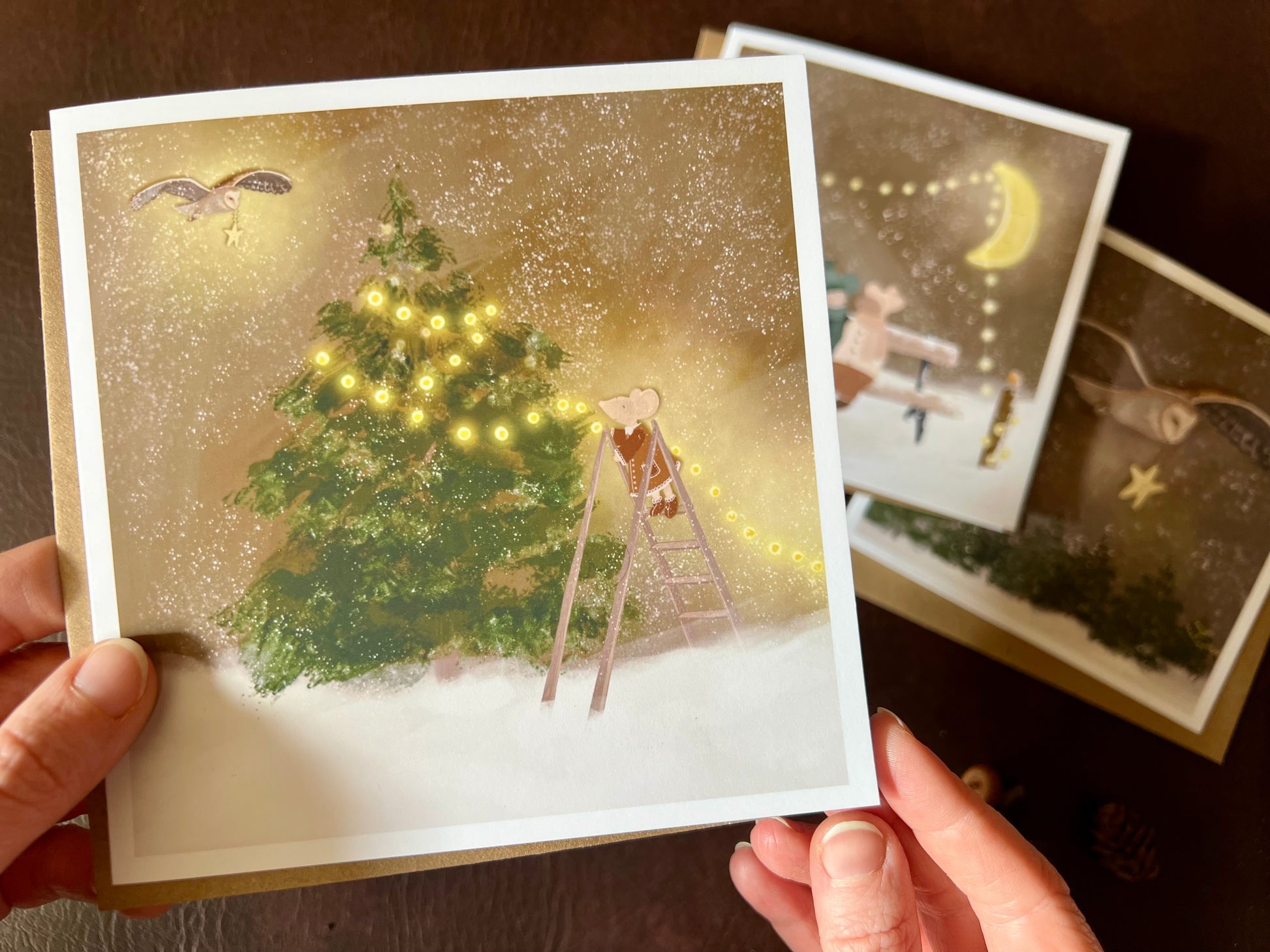 Premium Christmas card from a set of three woodland designs, featuring a storybook-style illustration of a mouse decorating a Christmas tree with fairy lights in a snowy winter scene. The card is shown in close-up being held in two hands, with the other two designs softly out of focus in the background. Ideal for festive gifting, animal lovers and rustic Christmas card sets. Printed on thick 312gsm fine art card by a UK independent artist.