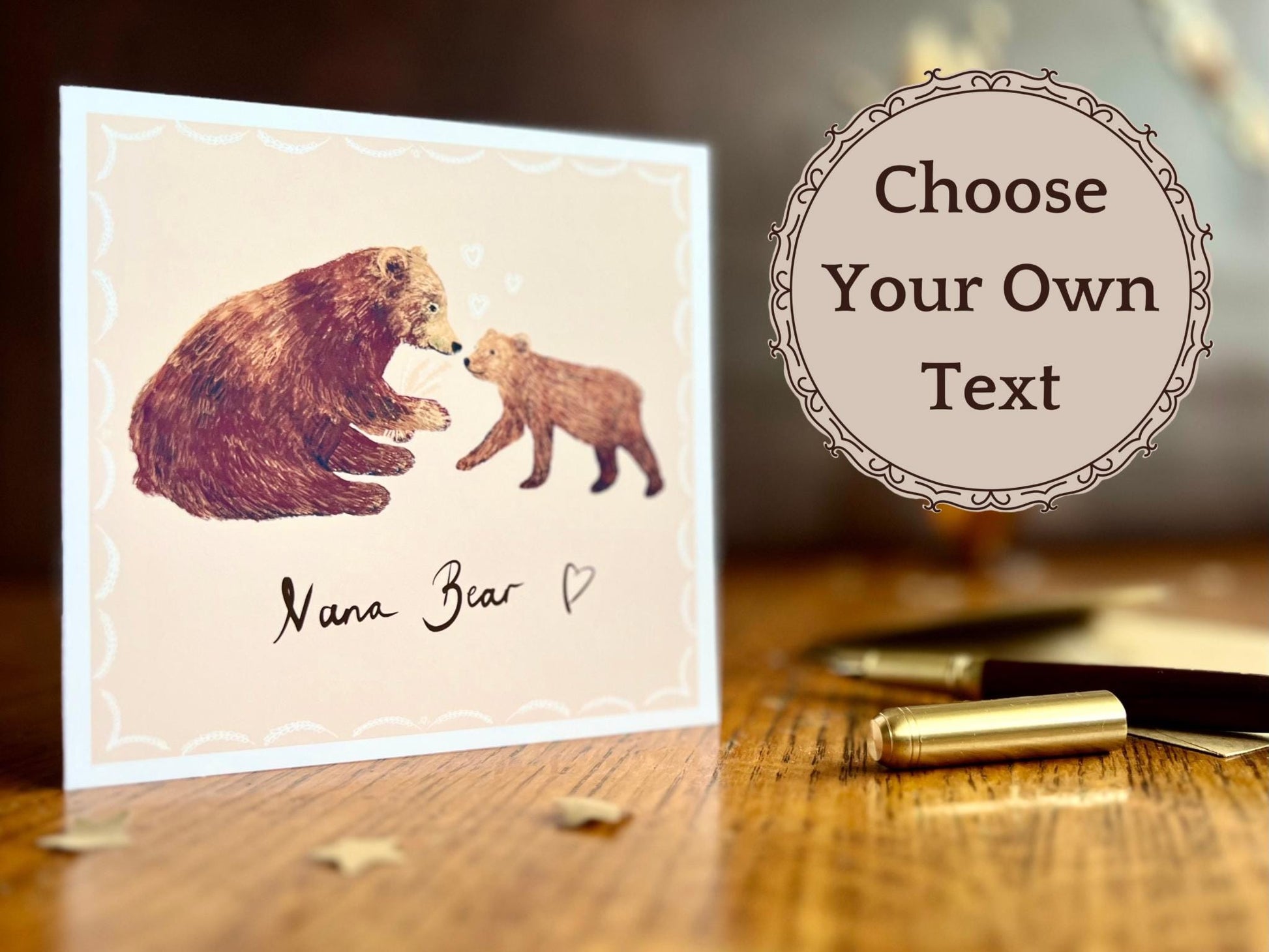 Lifestyle image of a handmade greeting card displayed on a tabletop featuring an illustration of a brown bear and cub with the text 'Nana Bear' on the front. A fountain pen sits in the foreground and a text overlay reads 'Choose Your Own Text', highlighting the card’s personalised options for different family names.