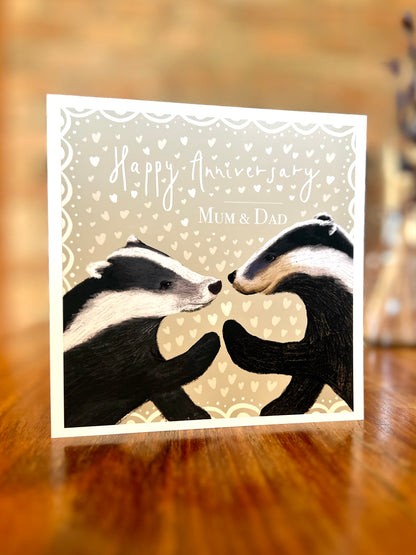 Personalised Happy Anniversary greeting card standing on a rustic tabletop in a farmhouse setting, featuring two black and white woodland badgers touching paw to paw on a pale sage green background with hearts and custom names, a premium high quality artist-designed art card.