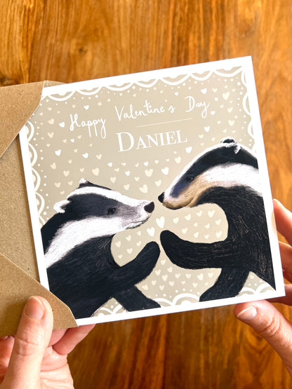 Personalised Valentine’s Day greeting card standing on a rustic tabletop, featuring two black and white woodland badgers touching paw to paw on a pale sage green background with subtle white hearts and custom name text.