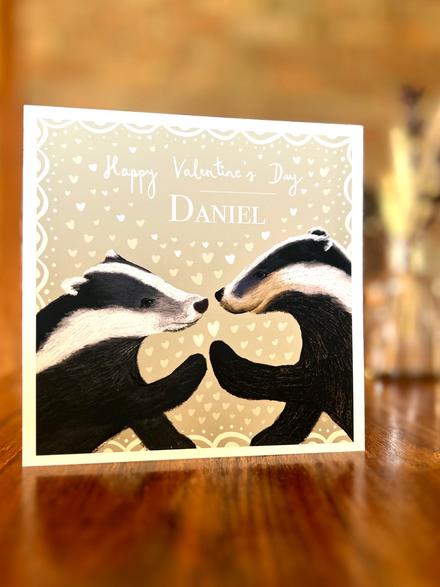 Personalised Valentine’s Day greeting card standing on a rustic wooden tabletop, featuring two black and white woodland badgers touching paw to paw on a pale sage green background with subtle white hearts and a custom name.