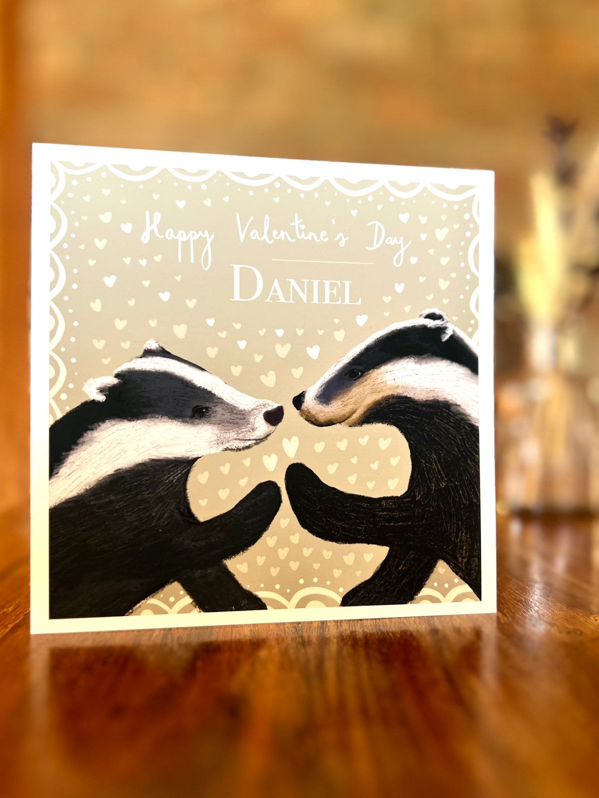 Personalised Valentine’s Day greeting card standing on a rustic wooden tabletop, featuring two black and white woodland badgers touching paw to paw on a pale sage green background with subtle white hearts and a custom name.