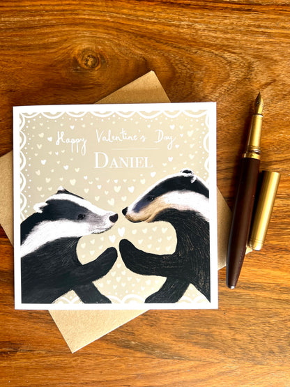Personalised Valentine’s Day greeting card with custom name laid flat on a rustic tabletop on top of a recycled fleck kraft envelope, featuring two black and white woodland badgers touching paw to paw, with a wooden fountain pen beside the card.