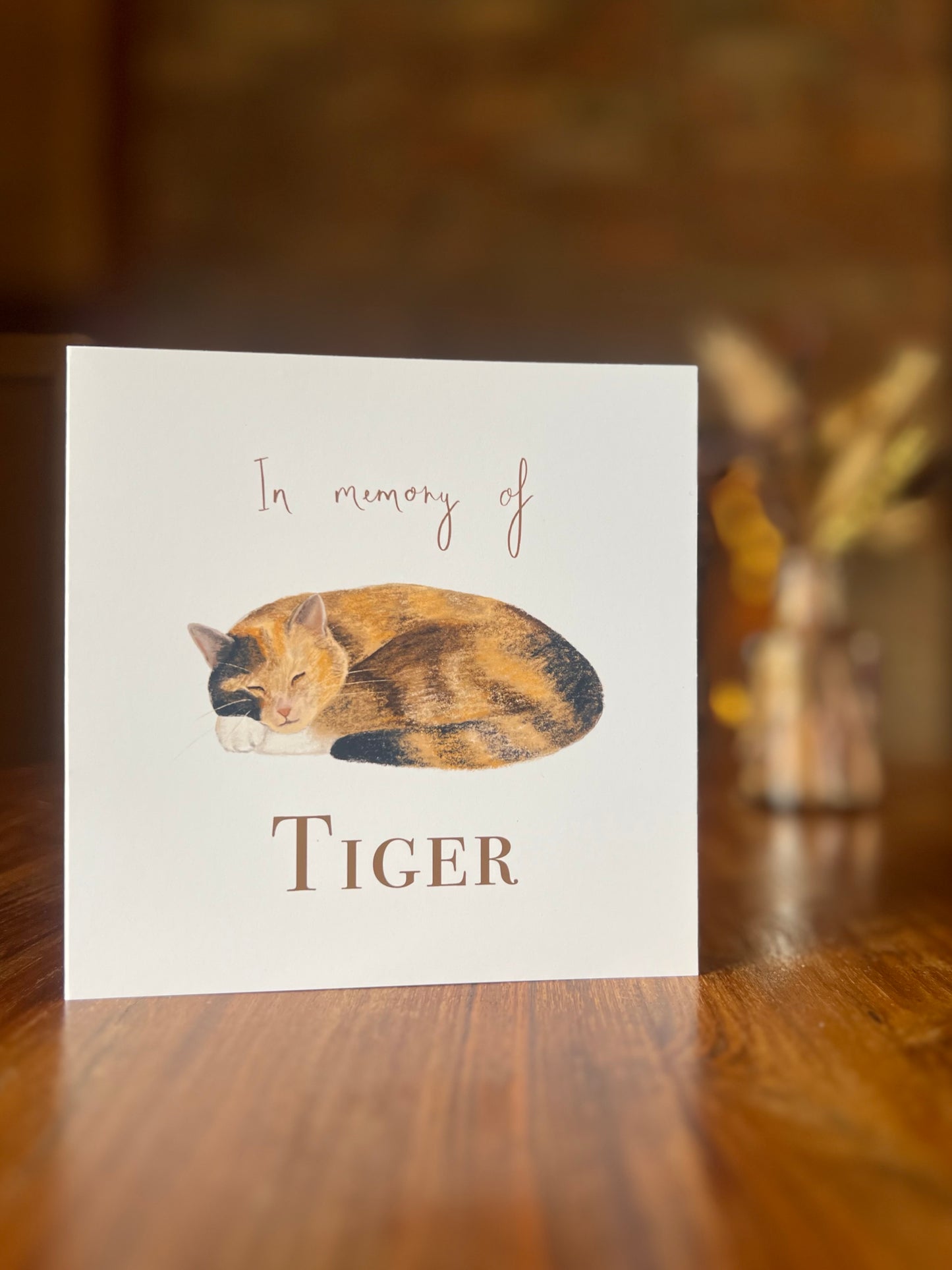 Personalised tortie cat memorial card displayed on a rustic wooden table, featuring a peaceful sleeping tortoiseshell and white cat illustration with a softly blurred background of dried flowers and a brick fireplace.