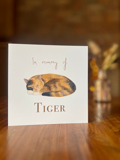Personalised tortie cat memorial card displayed on a rustic wooden table, featuring a peaceful sleeping tortoiseshell and white cat illustration with a softly blurred background of dried flowers and a brick fireplace.