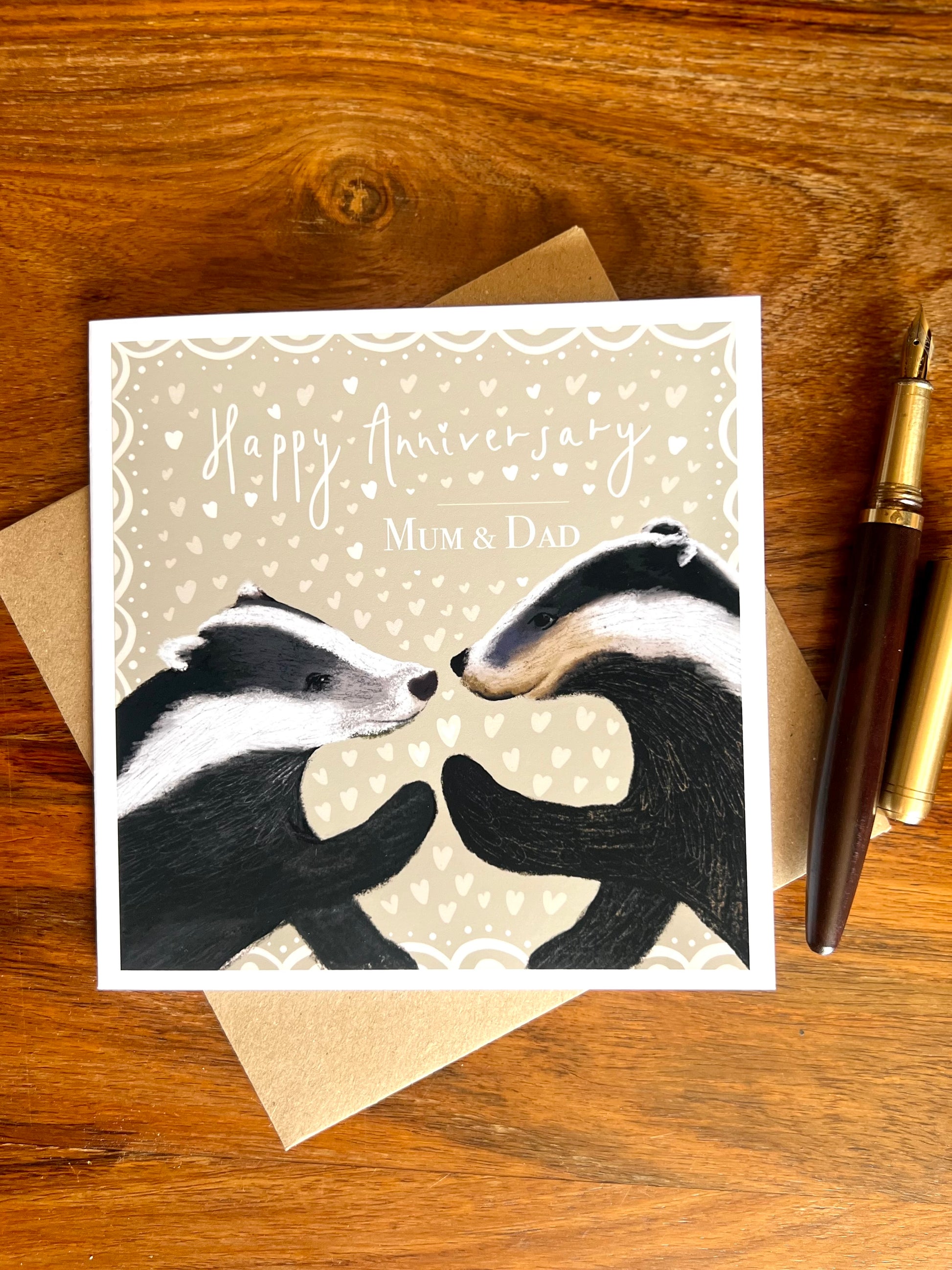 Personalised Happy Anniversary Mum & Dad greeting card laid flat on a wooden tabletop on top of a recycled kraft envelope, featuring two black and white woodland badgers touching paw to paw on a pale sage green background with hearts, alongside a wooden fountain pen, premium high quality artist-designed art card.