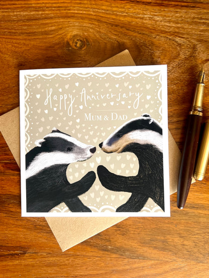 Personalised Happy Anniversary Mum & Dad greeting card laid flat on a wooden tabletop on top of a recycled kraft envelope, featuring two black and white woodland badgers touching paw to paw on a pale sage green background with hearts, alongside a wooden fountain pen, premium high quality artist-designed art card.