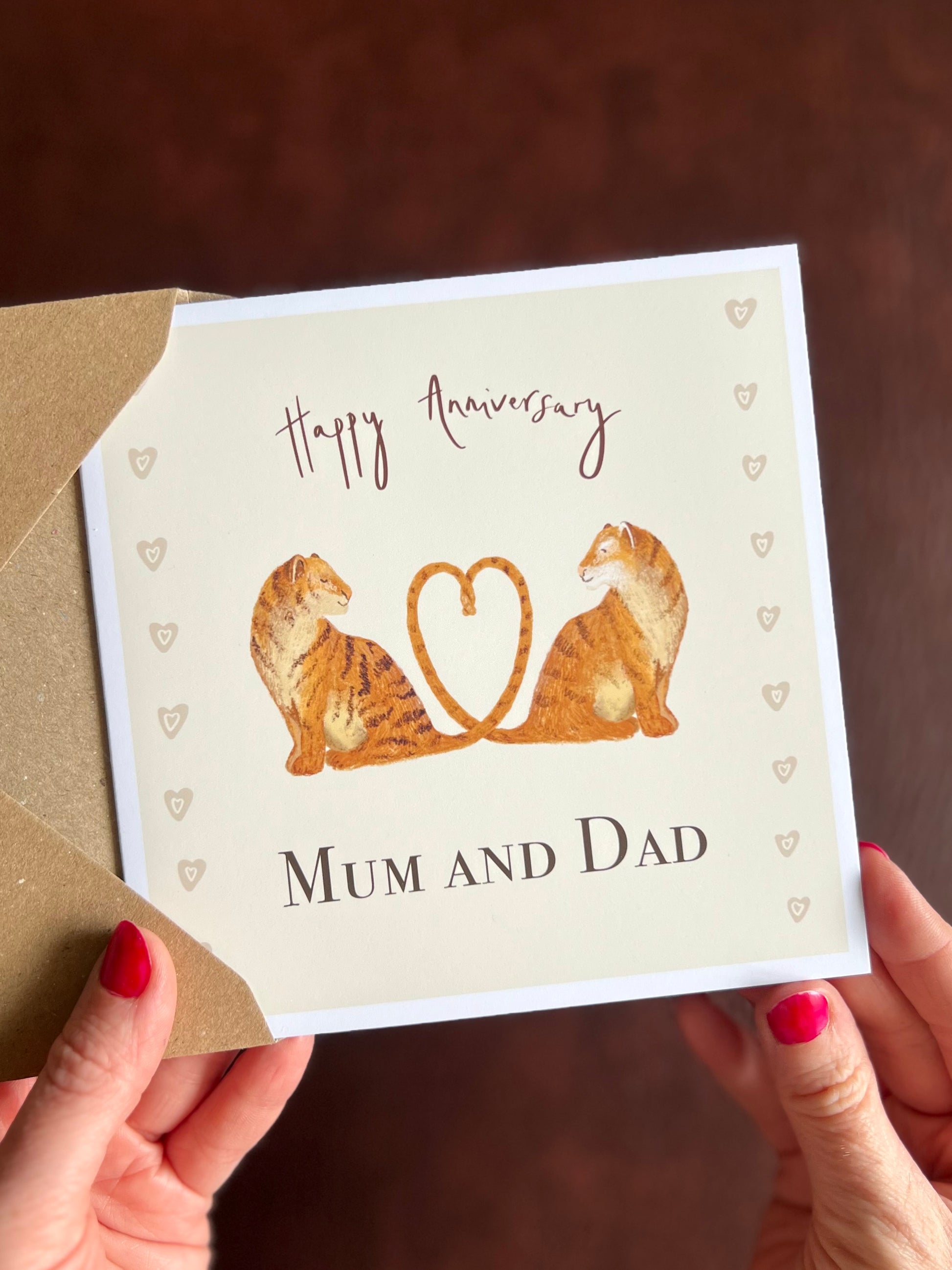 Hands holding a personalised tiger anniversary card featuring two illustrated tigers with tails forming a heart, with the text Happy Anniversary Mum and Dad in handwritten style font