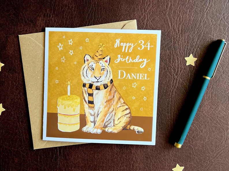 Premium personalised birthday card featuring an illustrated tiger wearing a black and amber striped birthday hat and matching scarf against an orange and amber background with birthday stars. The square card with a white border is shown laid flat, demonstrating custom name and age personalisation such as ‘Happy 34th Birthday, [Custom Name]’. Ideal for sports fans of teams nicknamed The Tigers, milestone birthdays and unique personalised birthday celebrations. Printed on thick 312gsm fine art card UK artist