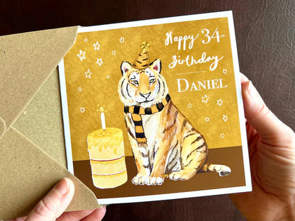 High quality personalised birthday card featuring an illustrated tiger wearing a black and amber striped birthday hat and matching scarf against an orange and amber background with birthday stars. The square card has a white border and is shown being held in two hands as it is removed from its envelope. Example text reads ‘Happy 34th Birthday, [Custom Name]’, demonstrating custom name and age personalisation. Ideal for sports fans of teams nicknamed The Tigers, milestone birthdays and special birthdays.