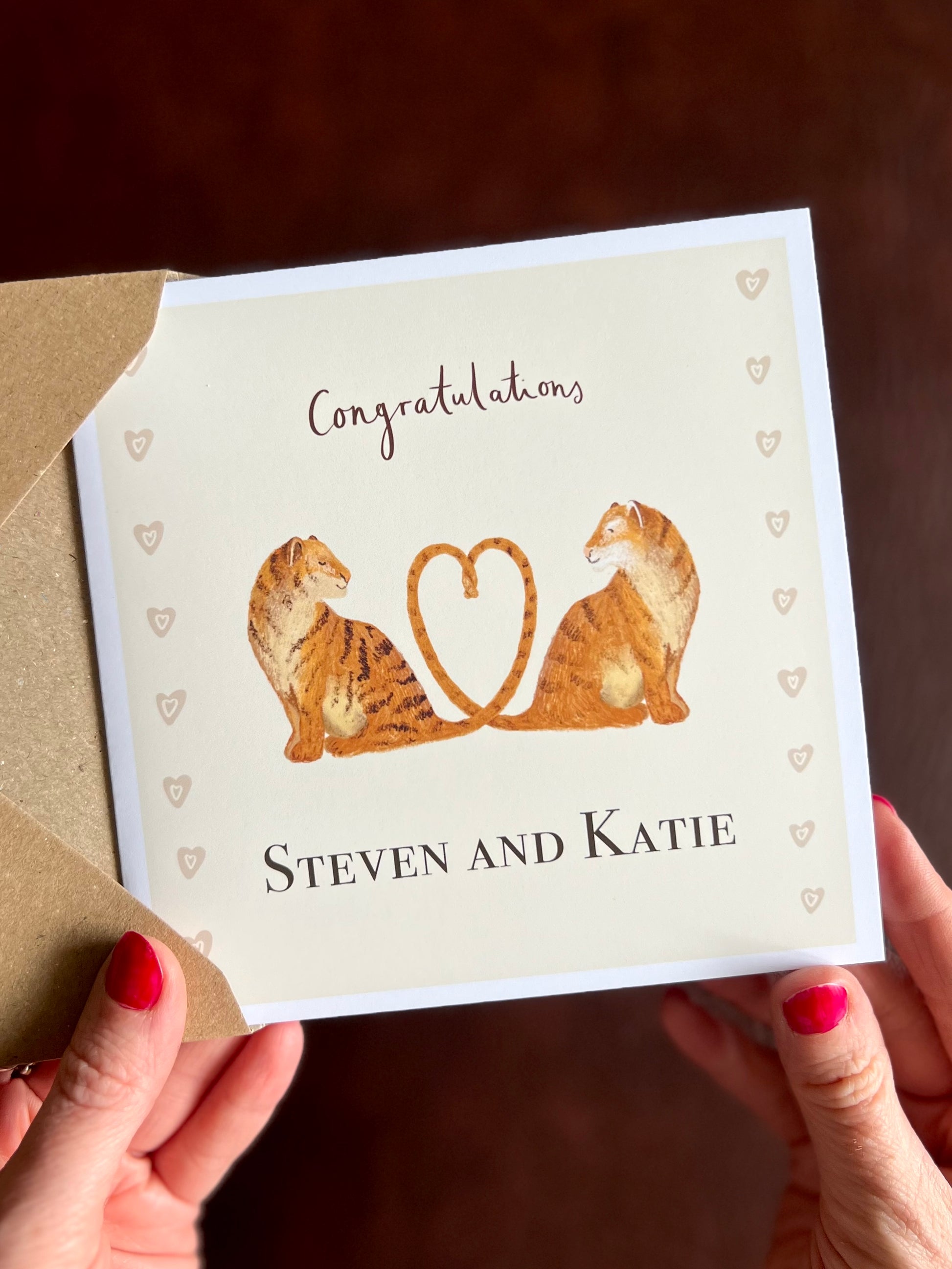Hands removing a personalised tiger engagement card from a kraft envelope featuring two illustrated tigers with tails forming a heart and custom couple names