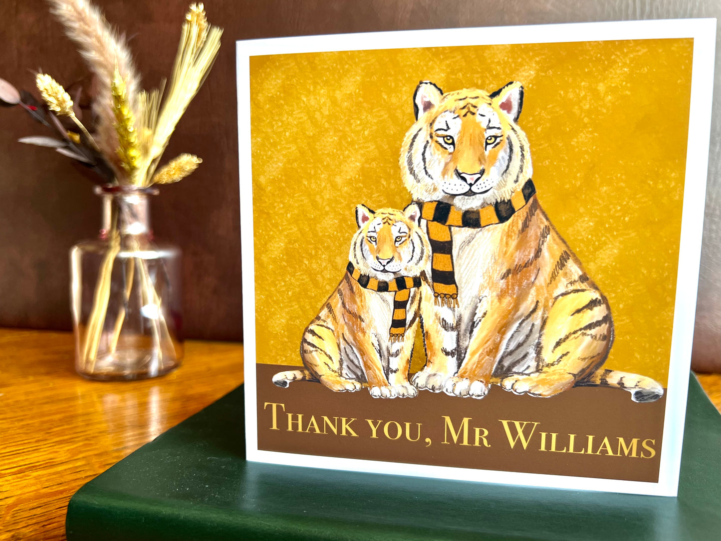 Premium personalised thank you card featuring illustrated tigers wearing black and amber scarves against a dark brown and burnt orange background. The square card is shown standing on a green leatherette book and displays the customisable text ‘Thank you, [Custom Name]’. Ideal end of term teacher appreciation card for teachers, teaching assistants or school staff, and suitable for fans of teams nicknamed The Tigers. Printed on thick 312gsm fine art card by a UK independent artist.