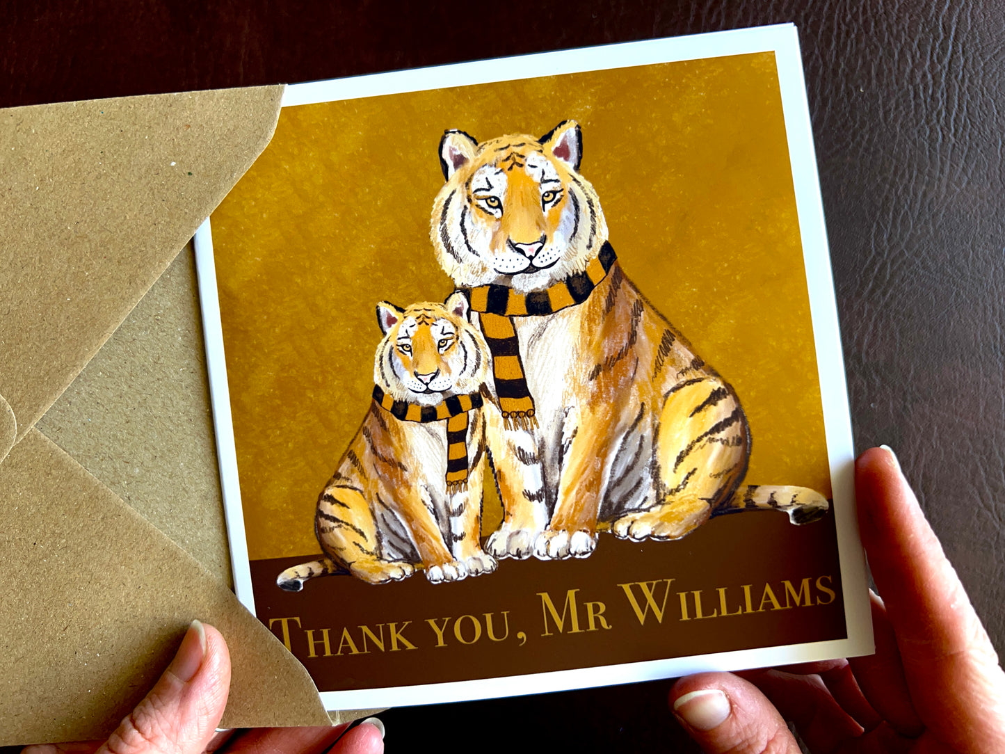 Premium personalised thank you teacher card featuring illustrated adult and younger tigers wearing black and amber scarves against a dark brown and burnt orange background. The square card is shown being held in two hands and displays the customisable text ‘Thank you, [Custom Name]’. Ideal end of term thank you card for teachers, teaching assistants or school staff, and suitable for fans of sports teams nicknamed The Tigers. Printed on thick 312gsm fine art card by a UK independent artist.