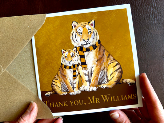 Premium personalised thank you teacher card featuring illustrated adult and younger tigers wearing black and amber scarves against a dark brown and burnt orange background. The square card is shown being held in two hands and displays the customisable text ‘Thank you, [Custom Name]’. Ideal end of term thank you card for teachers, teaching assistants or school staff, and suitable for fans of sports teams nicknamed The Tigers. Printed on thick 312gsm fine art card by a UK independent artist.