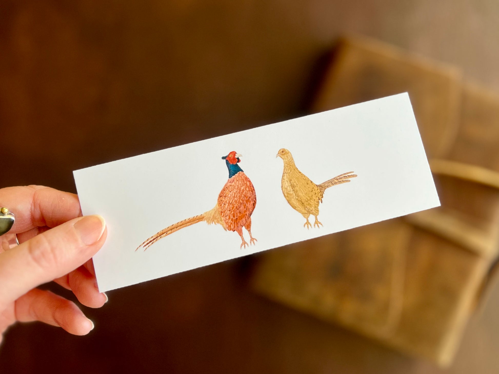Hand holding a handcrafted pheasant bookmark featuring a male and female pheasant illustration on a clean white background.