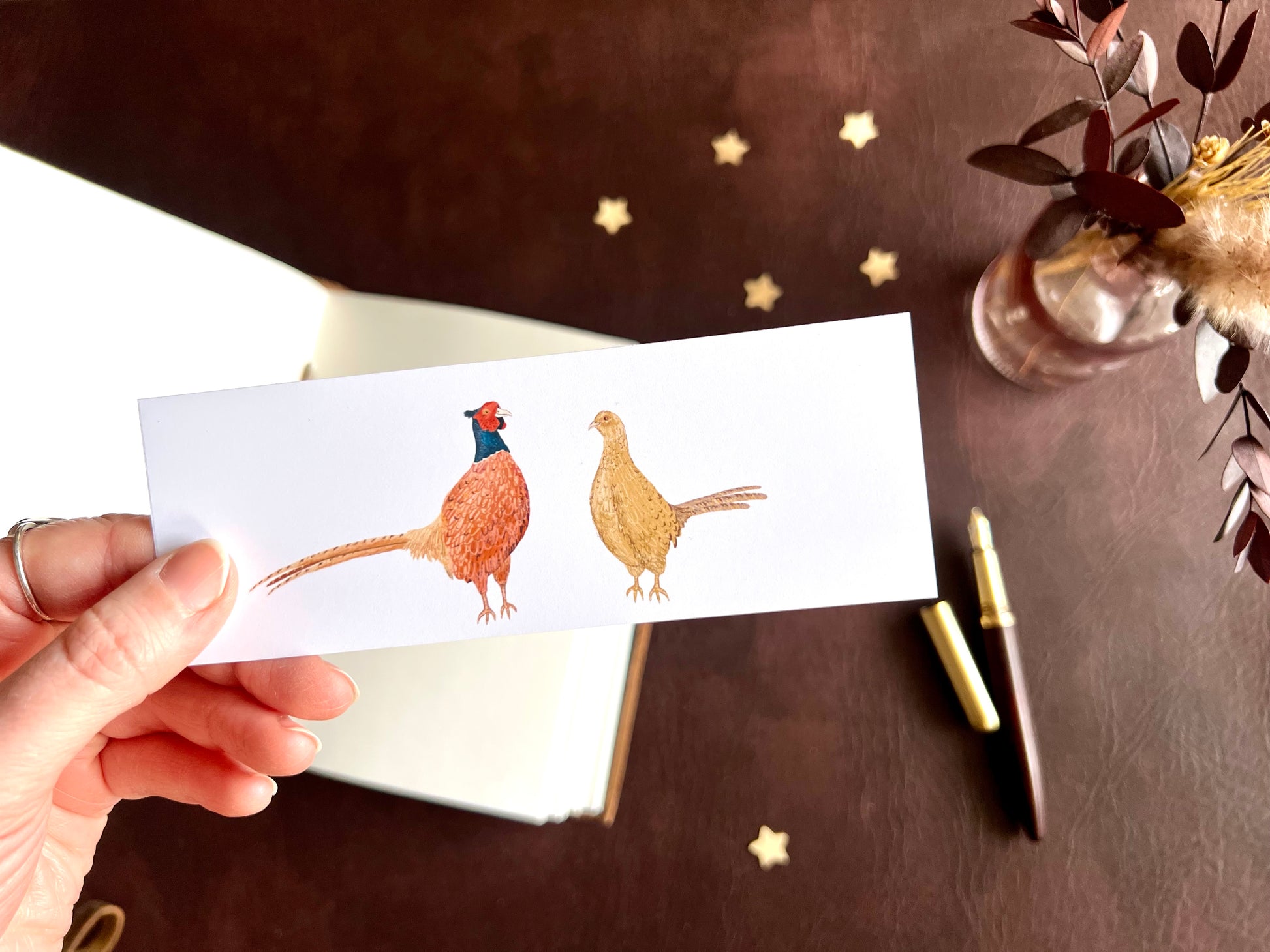 Pheasant bookmark shown with a fountain pen and open sketchbook on a cosy writing desk with a rustic British countryside inspired aesthetic.