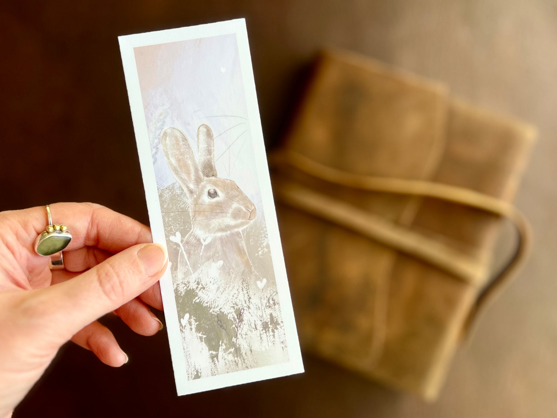 Hand holding a handcrafted rabbit bookmark featuring a rabbit illustration against a pale pink sky surrounded by subtle plants and flowers.