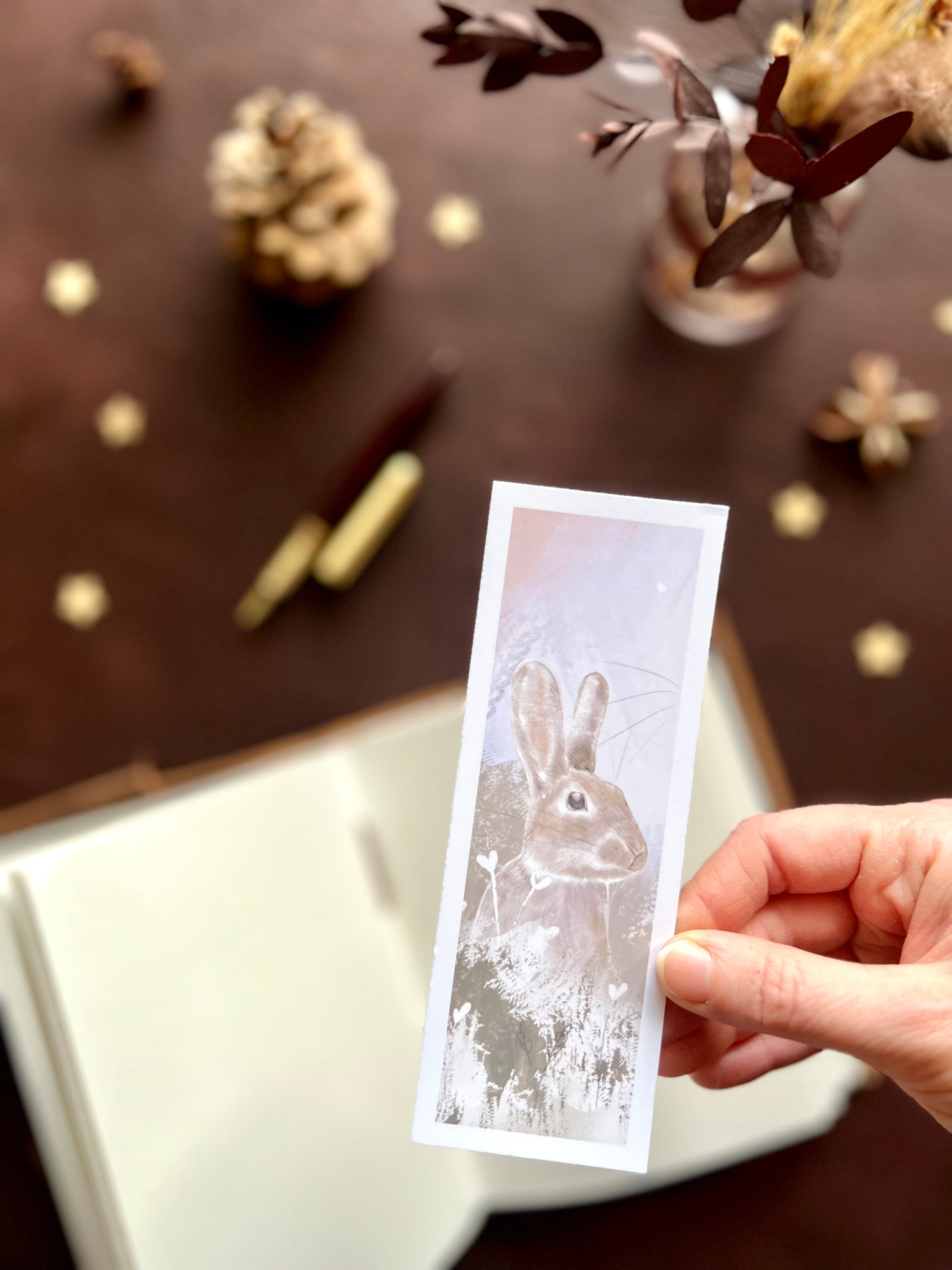 Close-up of a handcrafted rabbit bookmark featuring a pale pink sky illustration with subtle plants and flowers, photographed in a soft nature inspired setting.
