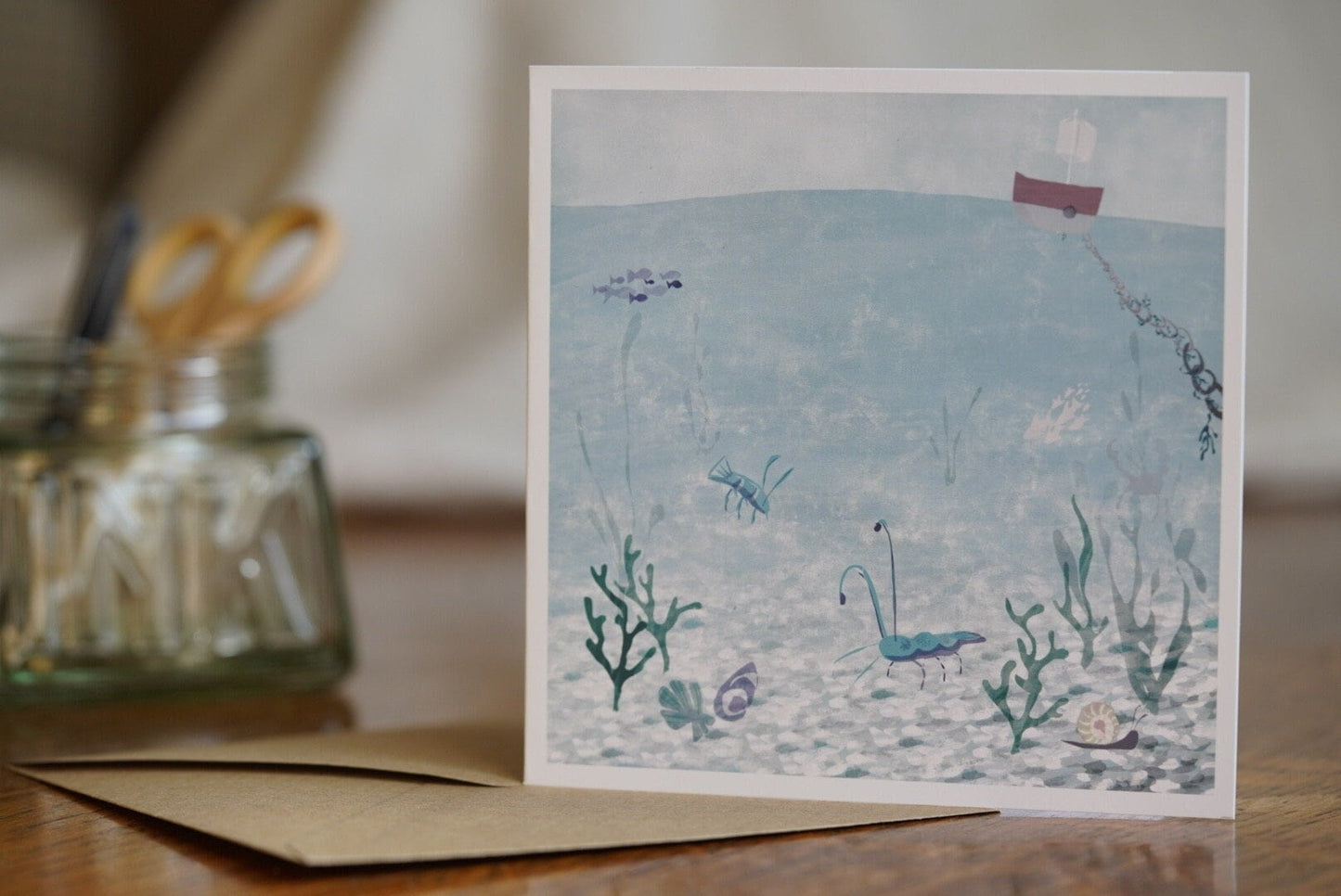Premium illustrated greeting card featuring a blue lobster and blue prawn in a crystal blue underwater coastal scene with a small boat above the water, displayed standing on a tabletop next to a recycled fleck kraft envelope. Joyful summery coastal art greeting card suitable for birthdays or keeping in touch.