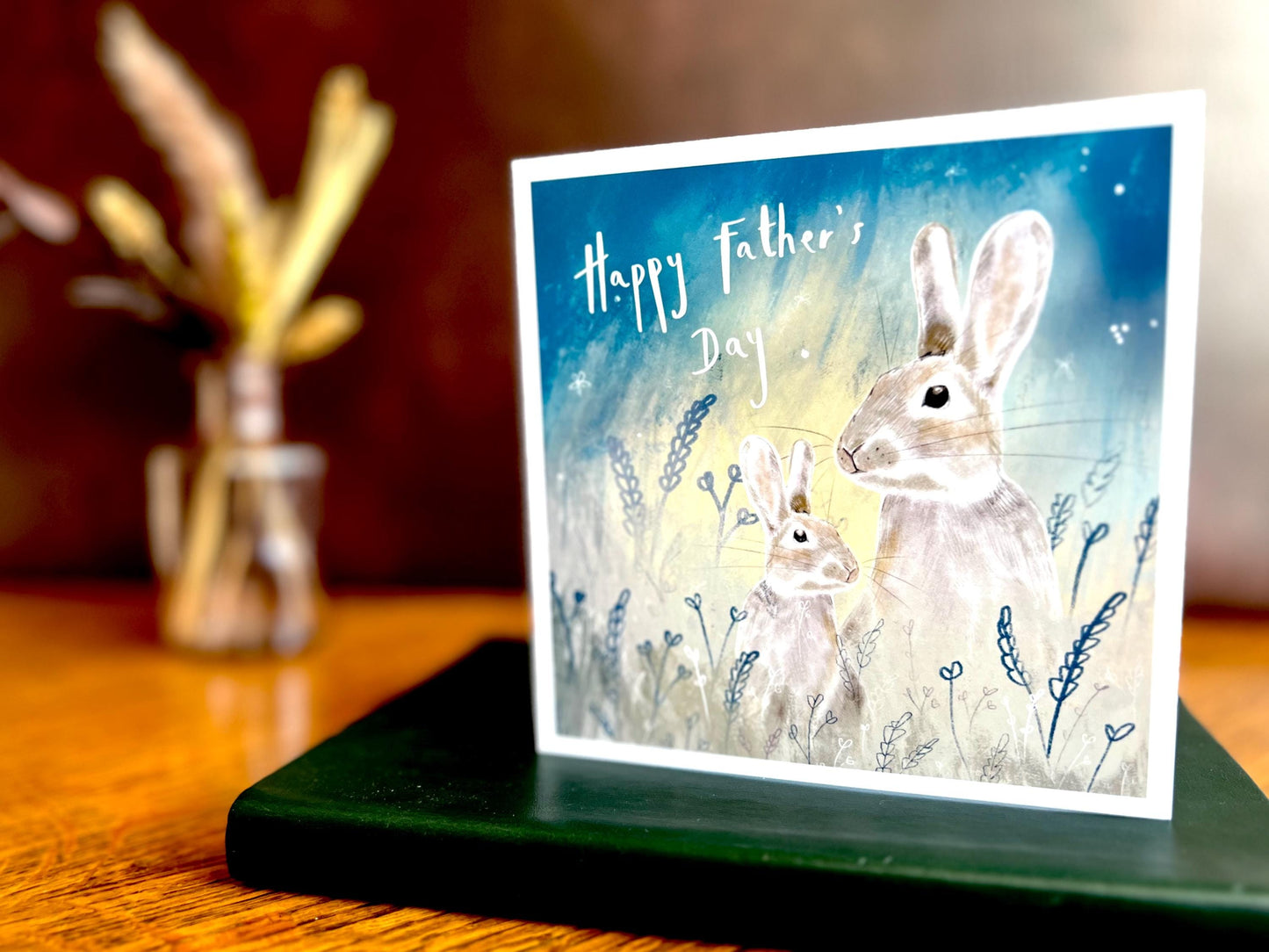 Premium Father’s Day greeting card featuring an illustrated father rabbit and young rabbit in a starry night sky design, styled standing on a green leatherette book placed on a wooden tabletop. The scene includes a brown leatherette backdrop and dried flowers in a vase in the background for a rustic lifestyle setting.