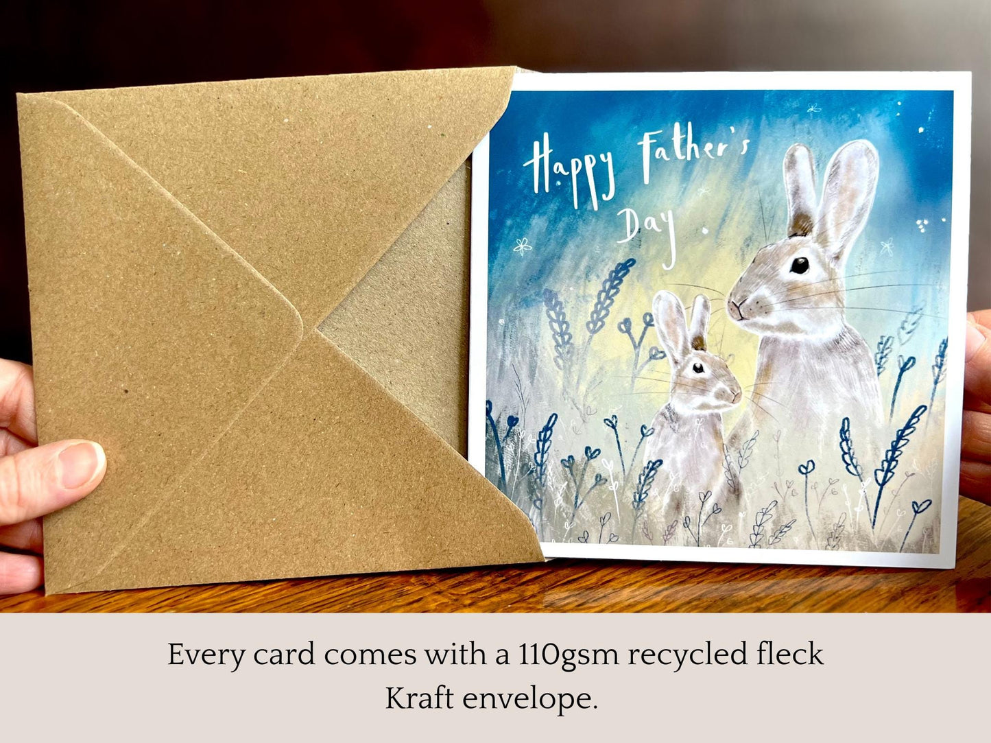 Handcrafted Father’s Day greeting card featuring an illustrated father and young rabbit design being removed from a recycled fleck kraft envelope. The card shows a starry night sky wildlife illustration with foliage and “Happy Father’s Day” text on the front. Premium greeting card unboxing scene.