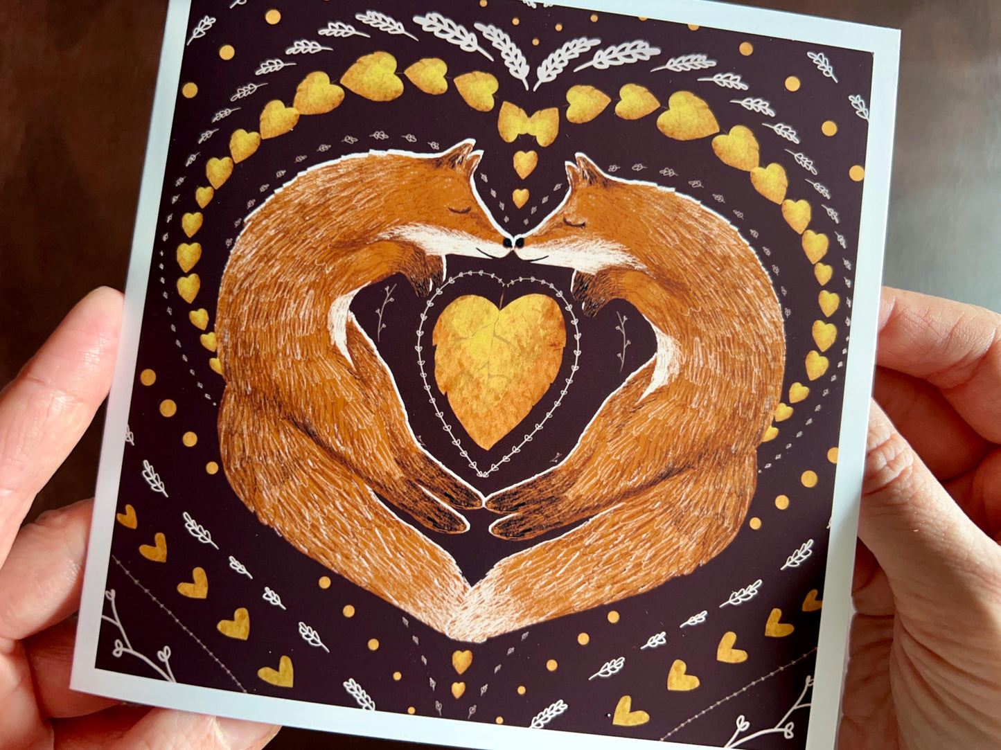 Premium illustrated greeting card featuring two foxes sleeping nose-to-nose with their bodies curled into a heart shape, surrounded by a symmetrical leaf and heart pattern on a deep maroon red background. Romantic love-themed card suitable for Valentine’s Day or anniversaries, shown in close up held in hands facing the camera.