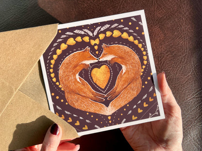 Premium illustrated greeting card featuring two foxes sleeping nose-to-nose with their bodies curled into a heart shape, surrounded by a symmetrical leaf and heart pattern on a deep maroon red background. Romantic love-themed card suitable for Valentine’s Day or anniversaries, shown held in hands.
