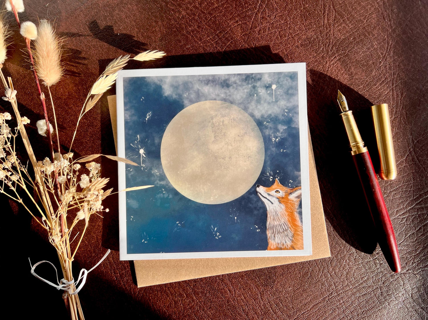 Premium illustrated greeting card featuring a fox looking up at a full moon in a starry night sky, laid flat on a recycled fleck kraft envelope on a brown leatherette tabletop. Styled flat lay includes a wooden fountain pen and dried flowers for a rustic lifestyle composition showcasing handcrafted design.