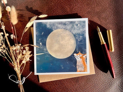 Premium illustrated greeting card featuring a fox looking up at a full moon in a starry night sky, laid flat on a recycled fleck kraft envelope on a brown leatherette tabletop. Styled flat lay includes a wooden fountain pen and dried flowers for a rustic lifestyle composition showcasing handcrafted design.