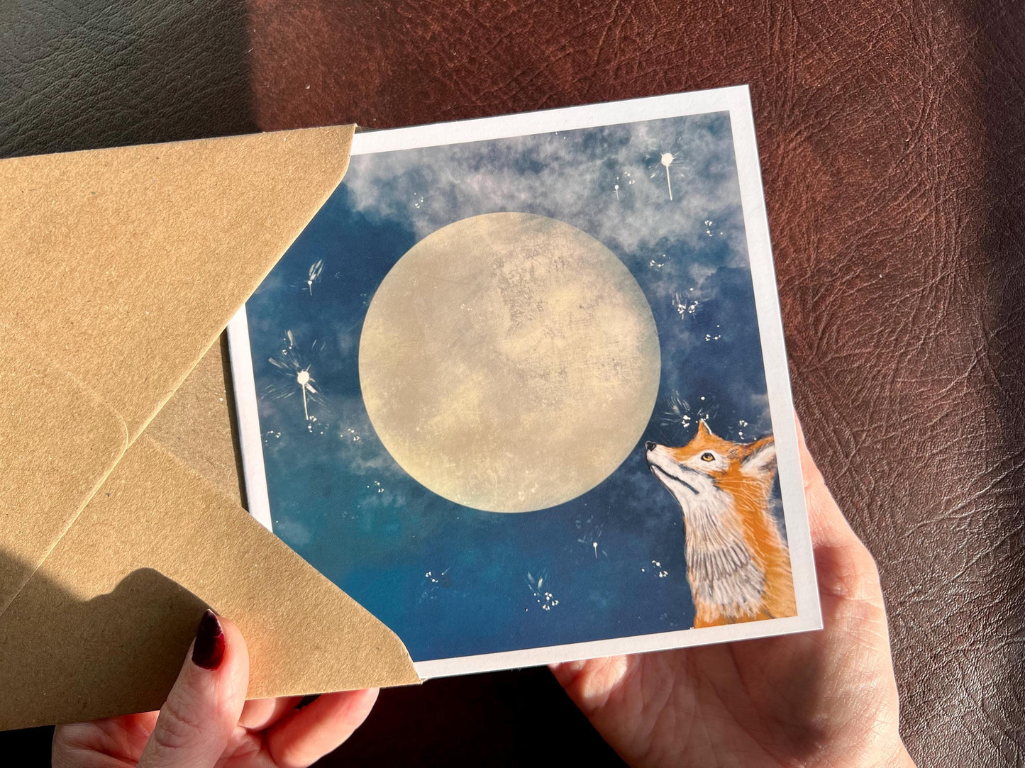 Premium illustrated greeting card featuring a fox looking up at a full moon in a blue starry night sky design, being removed from a recycled fleck kraft envelope and held in hands. Handcrafted wildlife art greeting card showcasing detailed magical, mystical illustration.