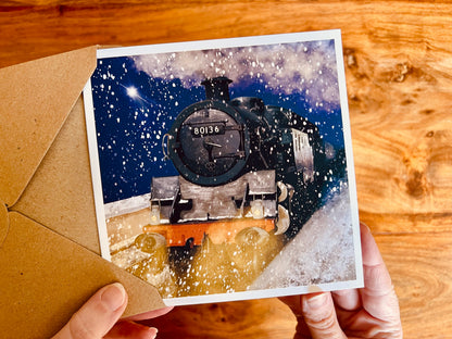 Hands holding a premium high quality special artisan greeting card being removed from a kraft envelope, featuring an illustration of steam train 80136 on a snowy night with falling snow, glowing headlights, a shining star in the sky and dramatic steam billowing through the scene.