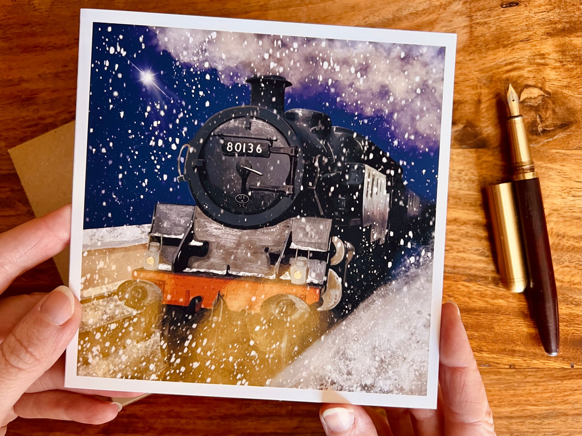 Aerial view of hands holding an artisan greeting card featuring steam train 80136 on a snowy winter night with falling snow, glowing headlights and a shining star, shown as the viewer looks closely at the design.