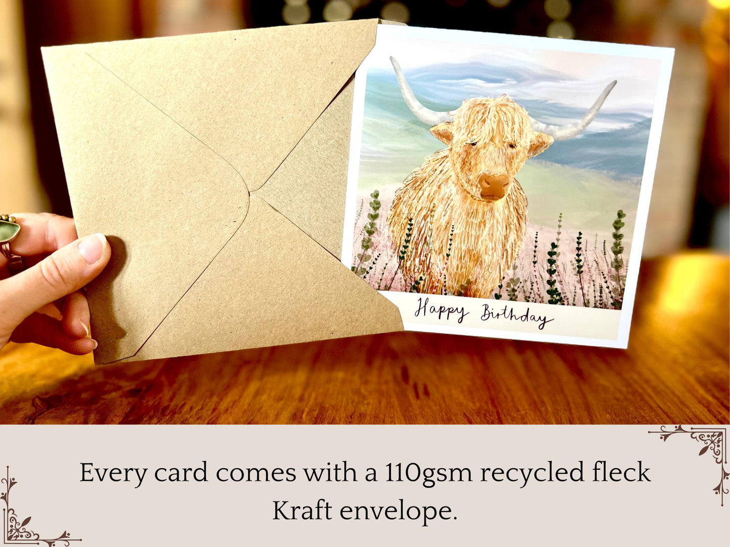 Premium illustrated birthday greeting card featuring a highland cow standing amongst heather in a tranquil countryside landscape, being removed from a recycled fleck kraft envelope. The image highlights that a 110gsm recycled fleck kraft envelope is included with each card, with “Happy Birthday” text beneath the illustration.