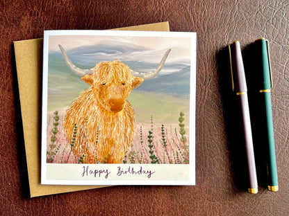 Premium illustrated birthday greeting card featuring a highland cow standing amongst heather in a tranquil countryside landscape, laid flat on a leatherette surface. The design includes the text “Happy Birthday” beneath the illustration.