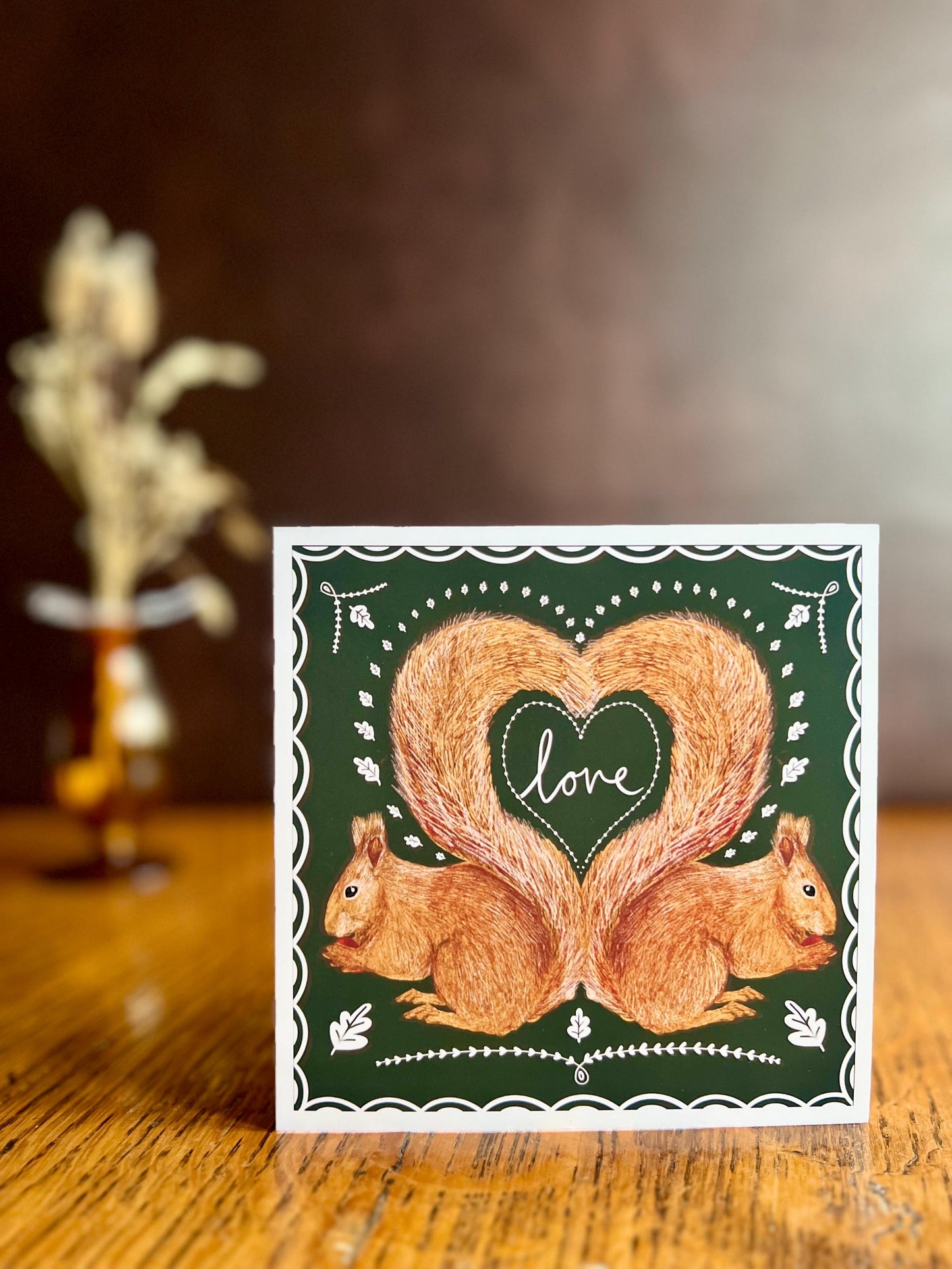 Premium illustrated greeting card featuring two red squirrels forming a heart shape with their tails and the word “love” in handwritten font, displayed standing upright on a wooden tabletop. Romantic Valentine’s Day and anniversary greeting card with dark green background and white scalloped border design.