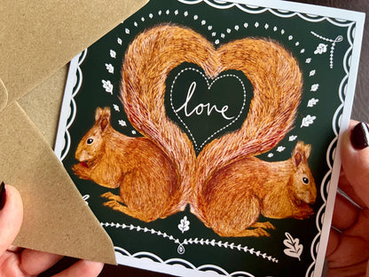 Premium illustrated greeting card featuring two red squirrels in a symmetrical design with tails forming a heart shape and the word “love” in handwritten font inside. The card has a white scalloped border and dark green background, shown being removed from a recycled fleck kraft envelope and held in hands. Romantic Valentine’s Day and anniversary greeting card design.