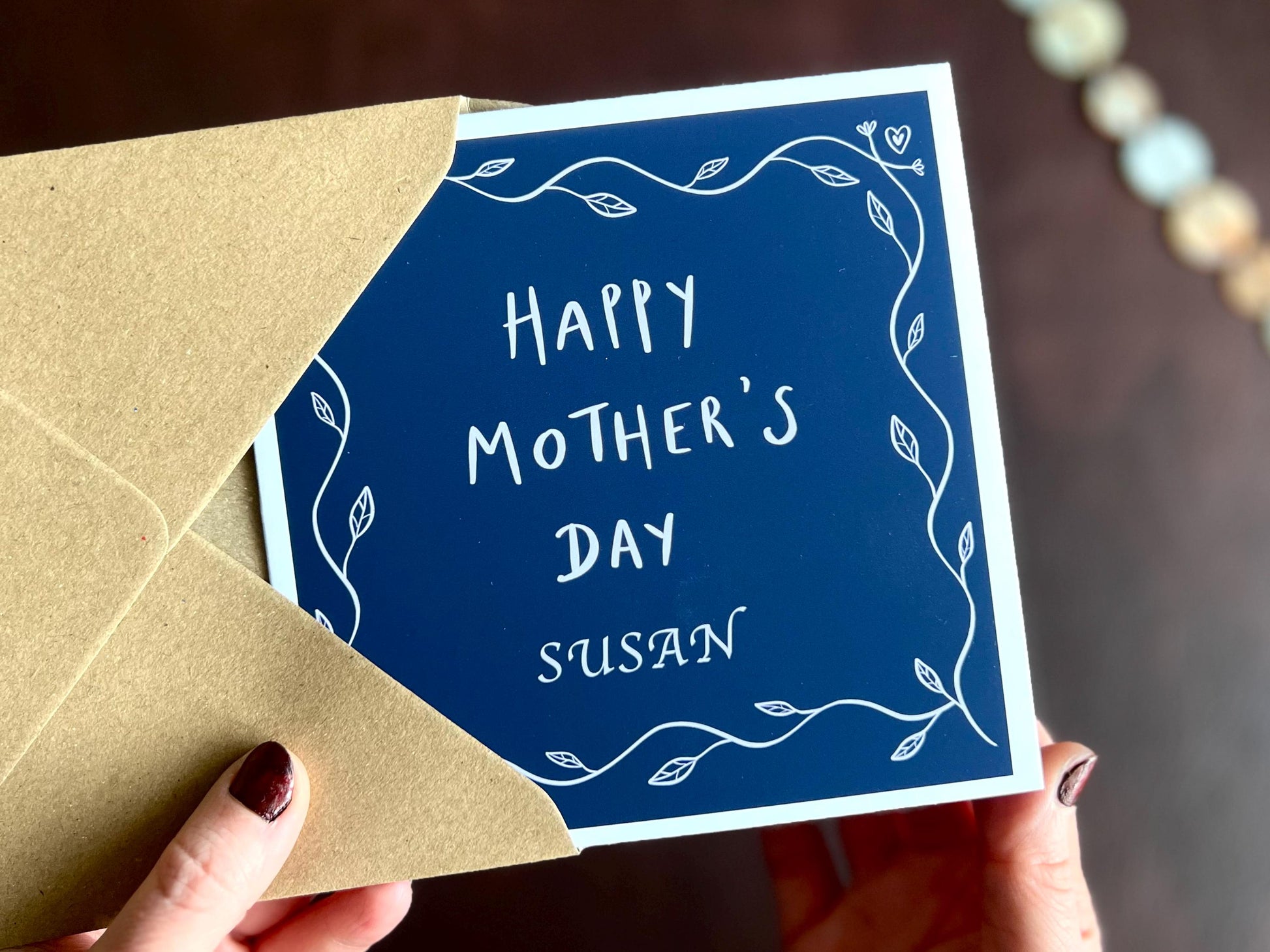 Premium Mother’s Day greeting card with a dark navy blue background and simple leaf border, featuring the text “Happy Mother’s Day” in the centre with optional personalisation such as “Mum”, “Step-Mum” or the name of a mother figure below. The card is shown being held in hands and removed from a recycled fleck kraft envelope.