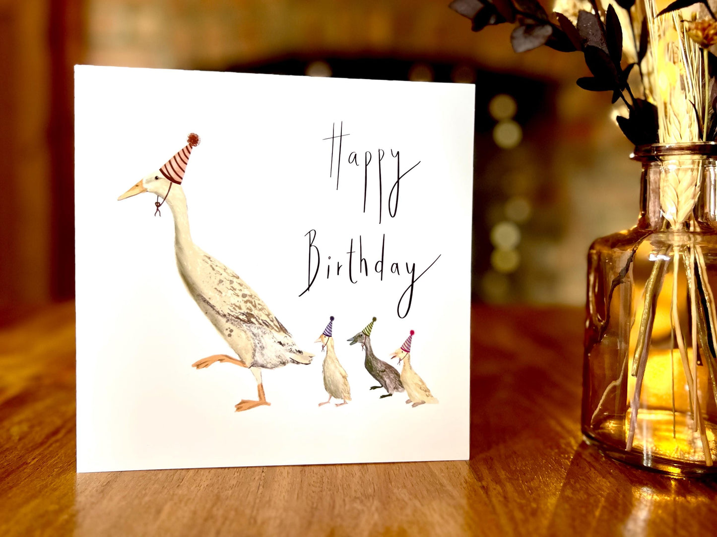 Premium illustrated birthday greeting card featuring a mother runner duck with three ducklings wearing party hats, displayed standing upright on a wooden tabletop. Playful countryside farm animal design with a plain white background and “Happy Birthday” text.